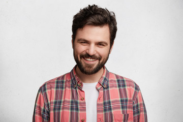 Pleased pleasant looking young student with beard and trendy hairdo wears casual checkered shirt, smiles pleasantly in camera, glad to pass session sucessfully, isolated over white background