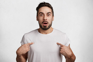 Scared frightened bearded male model wears casual white t shirt, indicates with fore fingers at blank space of clothes, being shocked with its price, isolated over white concrete wall. Unexpectedness