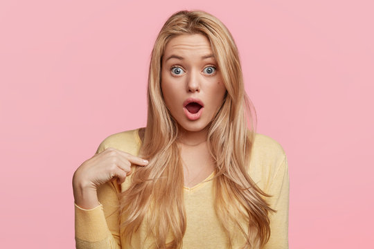 Portrait of beautiful female with terrified expression, looks with bugged eyes and keeps mouth opened, indicates at herself, isolated over pink background. Human reaction and emotions concept