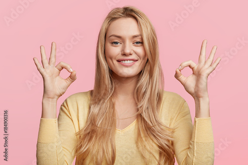 Fotografía Glad attractive woman shows ok sign as expresses approval, gestures in studio against pink background, has cheerful expression