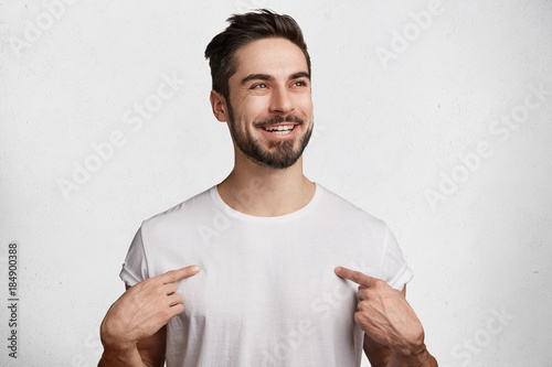 Fotografía Glad positive male with beard and mustache wears casual white t shirt, has shining happy eyes and pleasant broad smile, indicates at copy space for your design