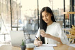 © bongkarn - Portrait of a young asian woman sitting at her desk while using her mobile phone.