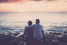 Gentlemen Couple Sitting At The Beach At Sunset. Hug And Love View From Back Rear. Foreverness Concept. People On Vacation And Ocean In Background