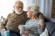 © Yakobchuk Olena - Honest conversation. Portrait of senior couple having dialogue at home while sitting on couch. Husband is cuddling wife while they holding hot cup of tea