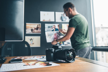 Aufkleber - Focus on camera and photos locating on desk in office. Side view unshaven serene man putting pictures on background. Creativity and occupation concept