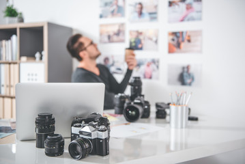Aufkleber - Focus on camera and notebook computer locating on table. Side view serene bearded man looking at photos on wall while tasting mug of coffee. Profession concept