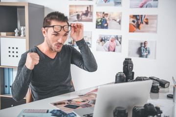 Aufkleber - Portrait of glad unshaven man flourishing arm while sitting at desk with laptop, gadgets and pictures in office. Happiness and occupation concept