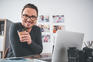 Aufkleber - Portrait of cheerful bearded young man working with photos and digital device while sitting at table in apartment. He tasting mug of tea. Break concept