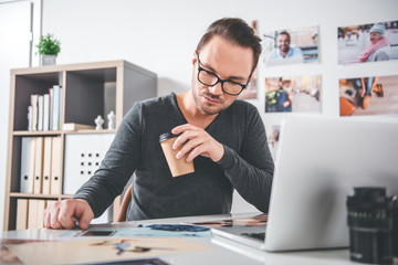 Aufkleber - Portrait of serene bearded man looking at different photos while drinking mug of appetizing beverage in room. Occupation concept