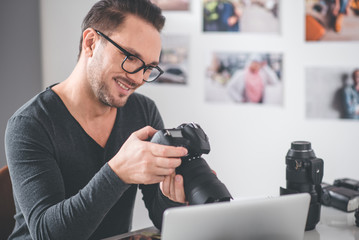 Aufkleber - Portrait of cheerful unshaven man choosing photos while keeping gadget for it. He situating at table in room. Labor concept