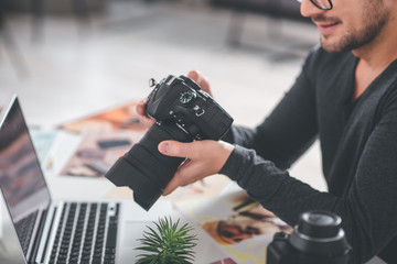 Aufkleber - Close up arms of cheerful bearded man holding professional camera. He looking at it while situating at desk with laptop. Job concept
