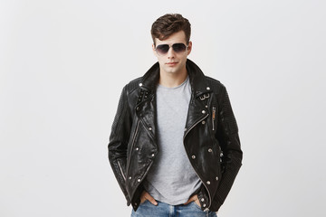 Wall Mural - Confident serious handsome man wears black leather jacket over gray t-shirt and stylish eyewear, looks directly into camera, isolated against gray background. People and style concept