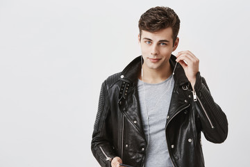 Wall Mural - Handsome positive man in black leather jacket with faint smile, listens music in earphones. Young caucasian male enjoys pleasant melodies, wearing white earphones, looking at camera with blue eyes.