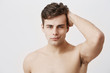 © Cookie Studio - Condifent young european man with stylish haircut and appealing eyes, being naked, touching dark hair, posing against gray background. Handsome male model with healthy clean skin gently smiling