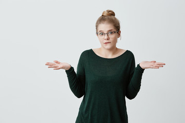 Clueless uncertain young woman with appealing appearance, blonde hair bun, shrugging shoulders, hesitates whether to go with friends on walk or study classes and preparing for exams. Life perception