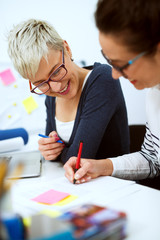 Close up of two smiling innovative stylish business middle aged women working together on problem solving while sitting in the office one next to another.