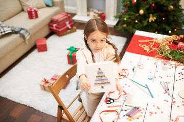 Wall Mural - Portrait shot of pretty little girl showing handmade Christmas card to camera while standing at wooden desk covered with confetti, pencils and wax crayons