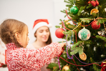 Wall Mural - Profile view of cute little girl wearing knitted sweater standing at Christmas tree and decorating it, her mother keeping eye on her