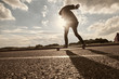 Back outdoor view of unrecognizable hipster guy sliding on skateboard along textured copy space asphalt. Skateboarder riding longboard on empty road in the morning. Flare sun, warm toned image