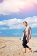 © raisondtre - Portrait of a cute confident elegant model girl posing barefoot in an elegant coat in a Sunny day on sandy beach on a blurred background of blue cloudy sky.