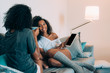 © DavidPrado - Happy young two black women sitting in the couch looking at a tablet and drinking coffee ...