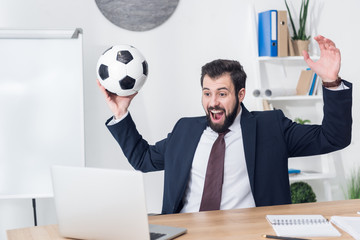 Wall Mural - excited businessman in suit with soccer ball looking at laptop screen at workplace in office