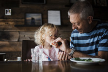 Grandfather Feeding Food To Granddaughter While Sitting At Table In Home