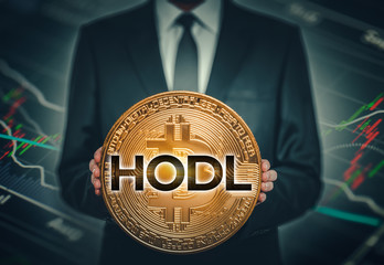 businessman holding big shiny bitcoin in hands in front of stock market data chart background and hodl word on bitcoin