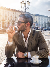 Handsome Young Man Drinking Espresso Coffee And Smoking Cigarette, Wearing Elegant Coat Posing At Table Outside On Urban Background.
