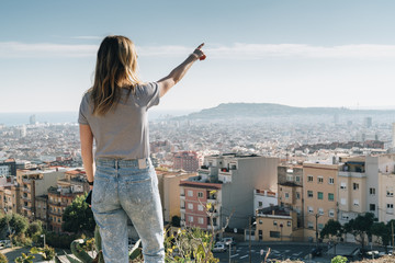 Wall Mural - Rear view of young woman wearing in stripped t-shirt standing on high point and looking at cityscape, pointing at something. Summer sunny day, rear view, bird's eye view of city, cityscape, horizon.