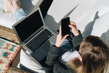 Wall Mural - Sunny day. View from above. Young businesswoman is sitting on white couch on terrace, using laptop and smartphone. Girl working, blogging, learning online. Distance work. Online marketing, education.