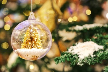 A little golden Christmas tree in the crystal clear bauble ball hangs as a decoration with blur background of Christmas tree with some snow on its leaf.