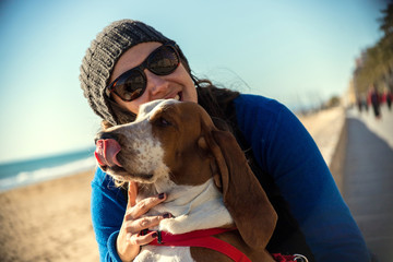 Woman loving her dog Basset Hound brown and white friend as she strolls along the edge of the beach in the winter sun with cap and blue jacket