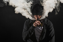 Young Bearded Man Wearing Cap Exhaling Smoke Of Electronic Cigarette Isolated On Black