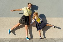 Two Girls Schoolgirl Elementary And High School, Posing In Front Of The Camera, On The Way To School. Background Gray Wall