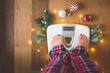 Top view of female feet in winter pajamas on digital scales or weight scale on wooden background surrounded with Christmas lights and decoration. Weight gain during holidays concept, vintage toned