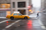 Fototapeta Nowy Jork - Panning motion image of a New York City yellow taxi cab in the snow as it passes through an intersection and past a pedestrian