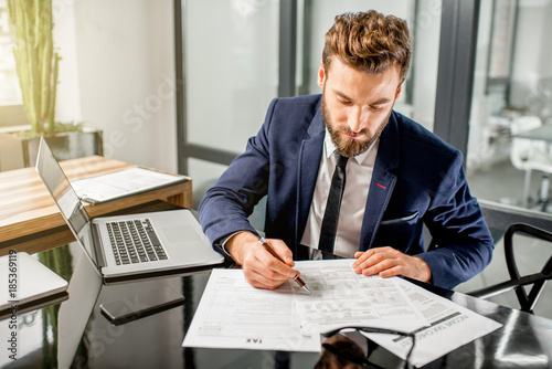 Cuadros en Lienzo Handsome tax manager dressed in the suit working with documents and laptop at th