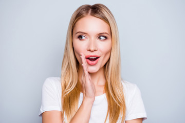 Don't tell anyone! Talkative mysterious pretty beautiful woman with blonde hair dressed in white tshirt is saying secret hot braking news and looking aside, isolated on grey background