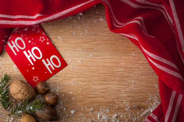 Merry Christmas and Happy holidays theme concept with wooden background, snow, fir tree, gift box card with the text Ho Ho Ho, mixed nuts and a red cloth, creating a border around the copy space