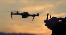 Close View Of Drone Pilot's Hands As He Controls Quadcopter Hovering Then Flying Off In The Background. Aircraft And Pilot Silhouetted Against Sunset Sky. Slow Motion Recorded At 60fps, 4K.