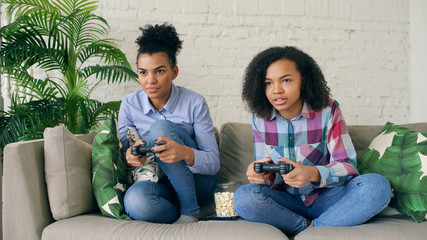 Wall Mural - Two mixed race curly girls sitting on couch play console computer games with gamepad and have fun at home