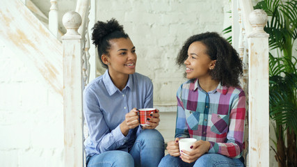 Wall Mural - Two african american curly girls sistres sitting on stairs have fun laughing and chatting together at home