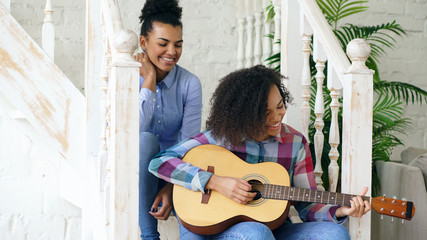 Wall Mural - Two brazilian curly girls sistres sitting on stairs and practice to play acoustic guitar. Friends have fun and singing at home