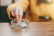 Focus On Caucasian Young Woman Hand Putting Out Cigarette On Glass Ashtray On Wooden Table, Cigarette Butt, Smoking Is Dying. Quit Smoking. Health Concept. Close Up Photo.