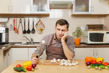 Sad upset caucasian young man in apron, brown shirt sitting at table with vegetable, cutting champignon mushrooms in light kitchen. Dieting concept. Healthy lifestyle. Cooking at home. Prepare food.