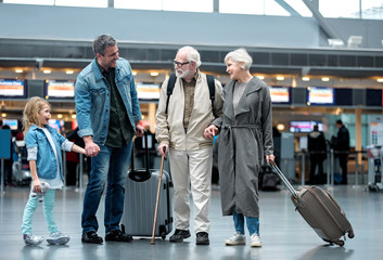 Unexpected meeting. Full length of cheerful aged couple with suitcases are standing at international airport with adult man and his little daughter. They are talking to each other with smile