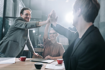 Aufkleber - Low angle happy males and cheerful woman rising hands into pyramid while locating at desk in room. Labor concept