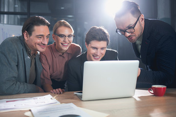 Portrait of happy female and glad men looking at laptop while locating at table in apartment. Occupation and business concept