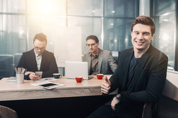 Poster - Everything is good. Portrait of happy young man demonstrating thumb up gesture. He sitting opposite comrades. Profession and communication concept